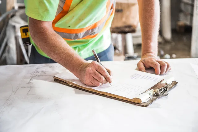 A construction worker writes notes on a clipboard at a construction site, emphasizing planning and precision. Anfahrtskosten und Einsatzgebiete vergleichen: Kriterien für regionale Dienstleister